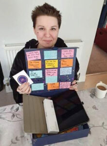 A woman is happily showing some things to the camera. She has short brown hair, wears a black hoodie. Some small tattoos are visible on her hands and fingers. In her hand is an opened gift box with loads of colourful messages glued to the top. Inside the gift box is a graphic tablet and a special pen to go with it.