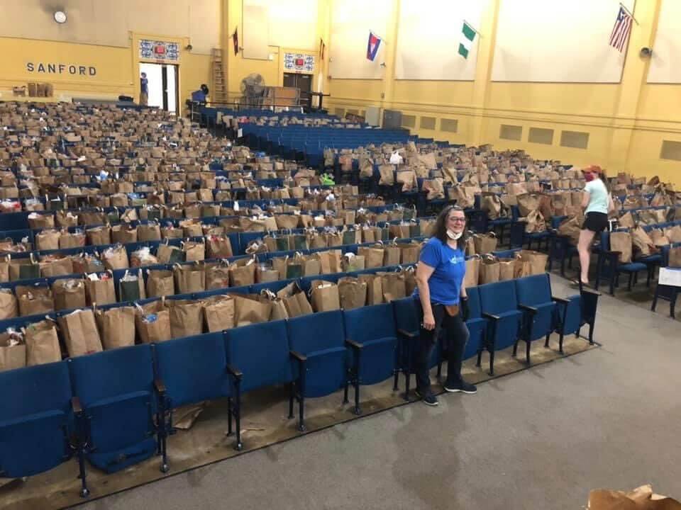 Auditorium with over 20 rows of seats each filled with a brown paper bag containing food from the food drive. Two volunteers in masks are filling bags. 