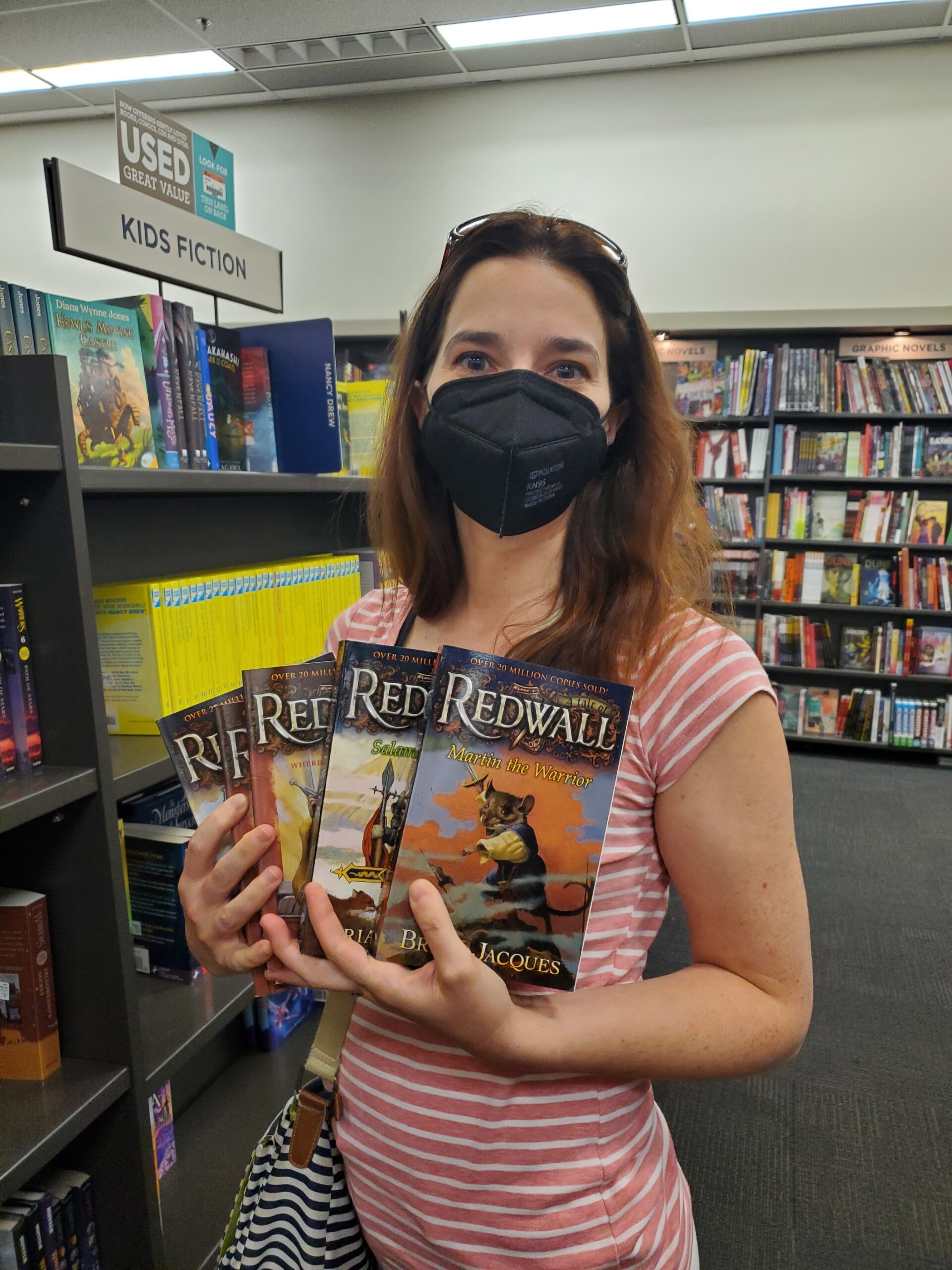 A young woman in a pink t-shirt is standing in a book shop. She has red-brown shoulder-length hair and is wearing a black medical mask. She is holding four fantasy books in her hands with the title "Redwall".