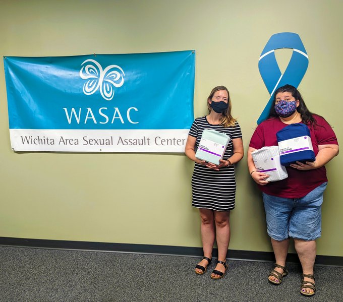Two women wearing face masks and holding sheet sets in front of a sign that says WASAC.