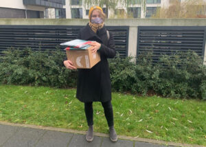 A woman in a black coat wearing a mask standing in front of a hospice center holding a box of donations