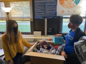 Two woman stand in a principal's office around a cardboard box that is filled with hats, socks, and gloves.