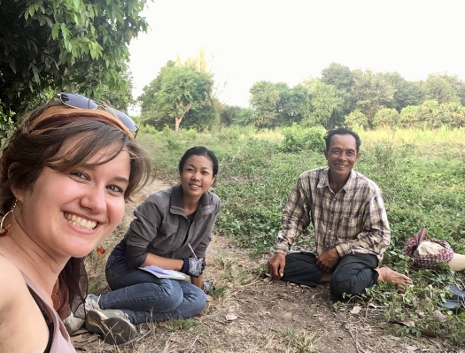 Christina smiling taking a selfie sitting in a field with a man and a woman in Cambodia