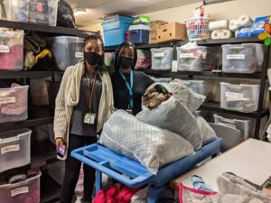 Two woman wearing face masks are standing next to each other in a room with many filled boxes in shelves. In front of the women lie white bin bags with coats on a table.