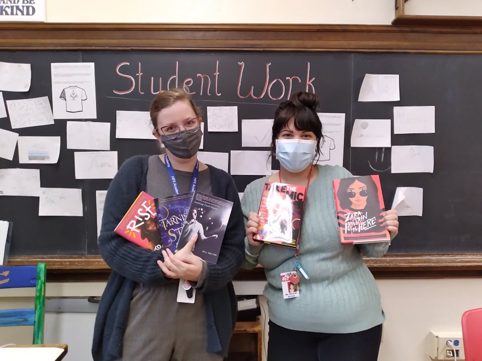 Two women stand in a class room. Behind them is a green board with slips of paper with drawings. Above that is chalk writing saying "Student Work". The two women both wear medical masks and hold books in their hands. One woman has dark hair in a bun and bangs and wears a blue shirt. The woman on the left wears a darker mask and darker clothes