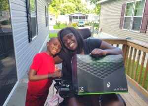Dominique who lives with quadriplegia, sits in her mobility device holding her new Xbox. Her young son stands next to her while they hug. 