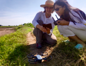 Christina crouched on a river bank with a Cambodian student and a drone