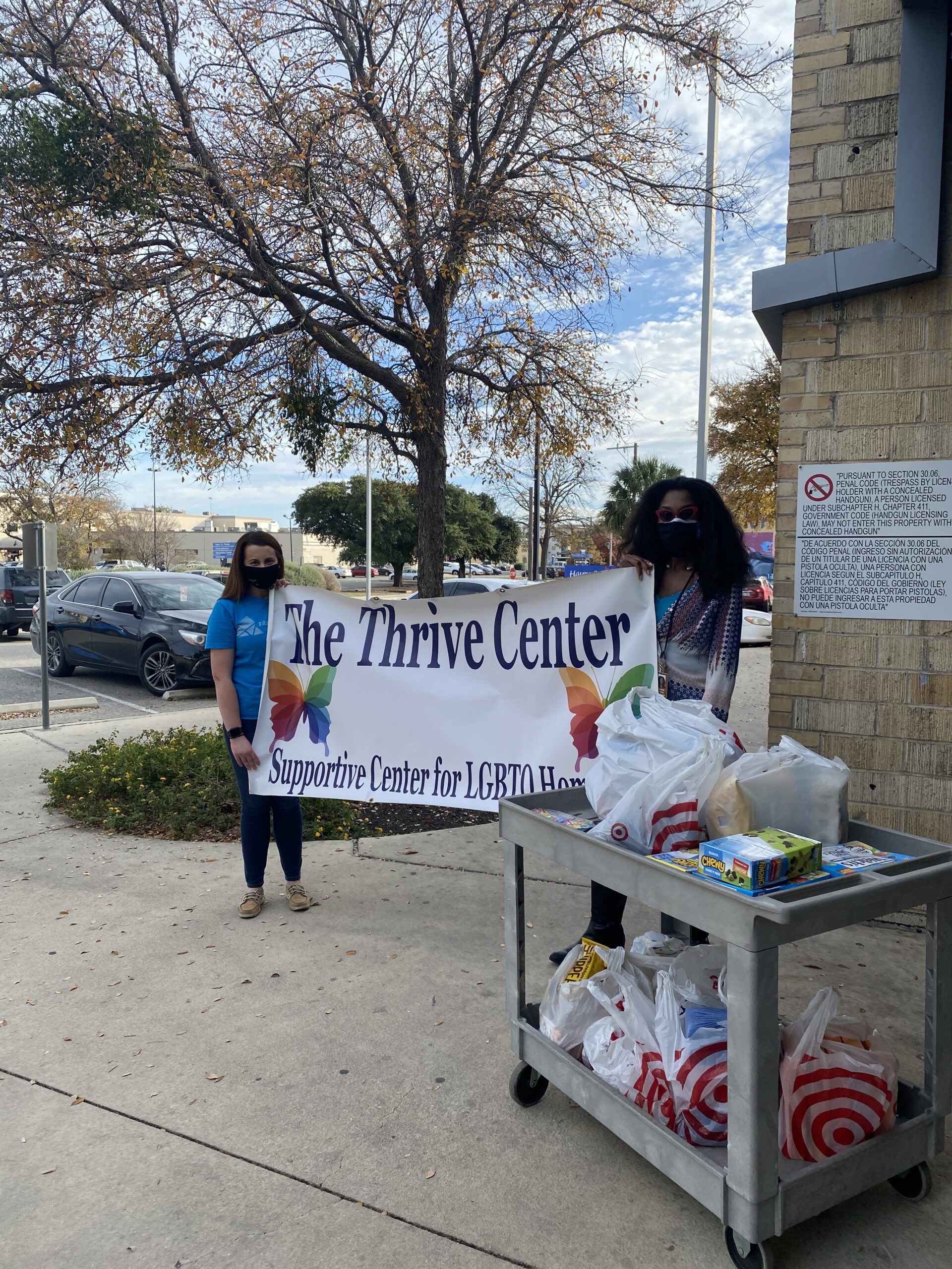 Two women stand outside holding a banner that reads "The Thrive Center" then "Supportive Center for LGBTQ" next to a cart with shopping bags full of items. 