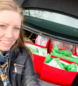 A woman stands in front of an open car trunk that has seven red gift bags inside.