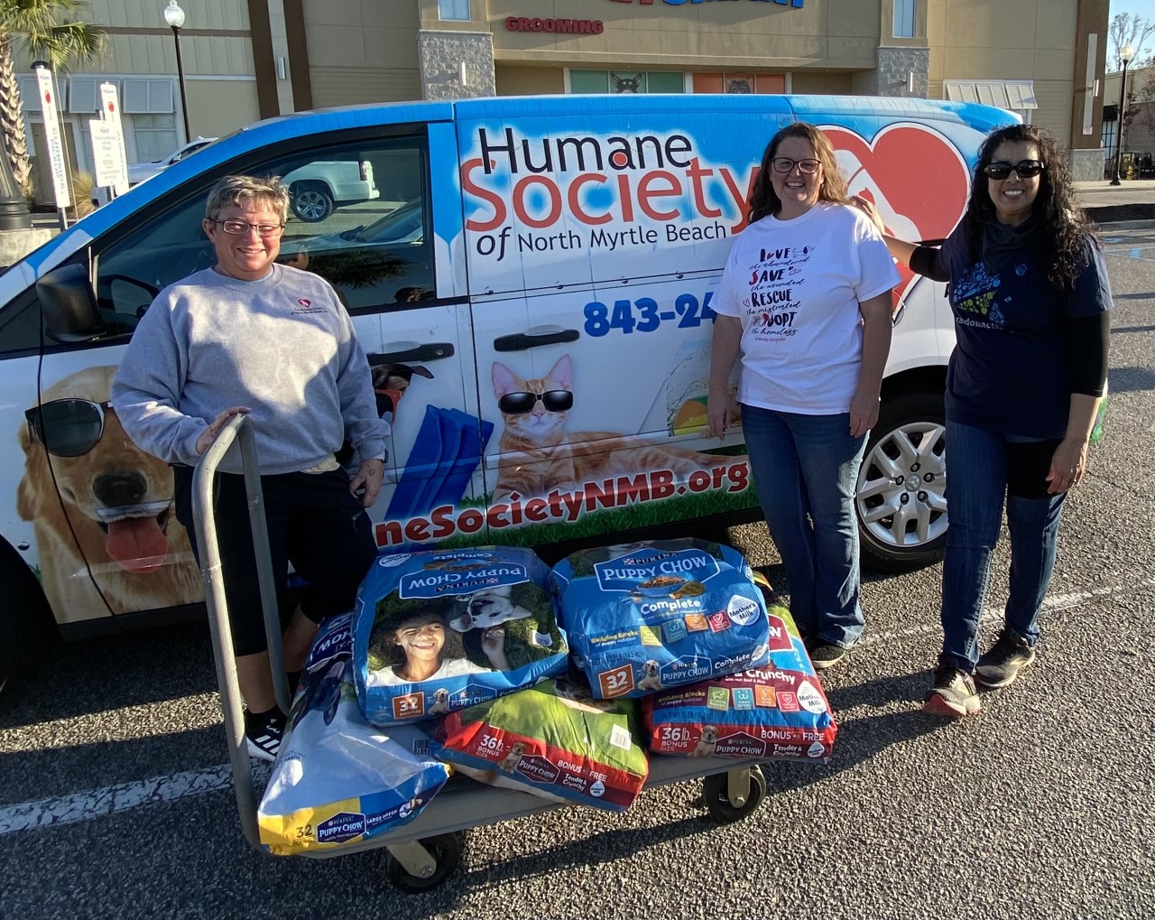 Three people stand in front of a blue Humane Society van with five large bags of puppy food.
