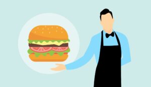 A man in a blue shirt, black bow tie, and black apron gestures to a large hamburger