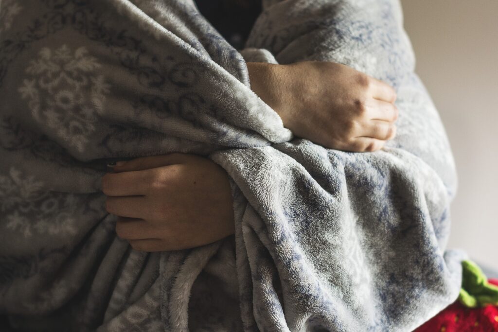 A part of a female body wrapped in a grey soft blanket. Hands are grabbing the blanket, pulling it close.