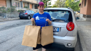 A woman with short, pink hair wearing a blue Random Acts shirt holds up three brown gift bags