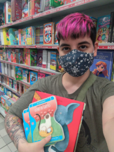 A woman with short, pink hair wearing a mask and holding kids books