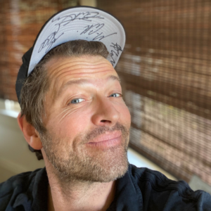 Misha smiling and wearing a baseball cap showing the underside of the rim featuring signatures of several people